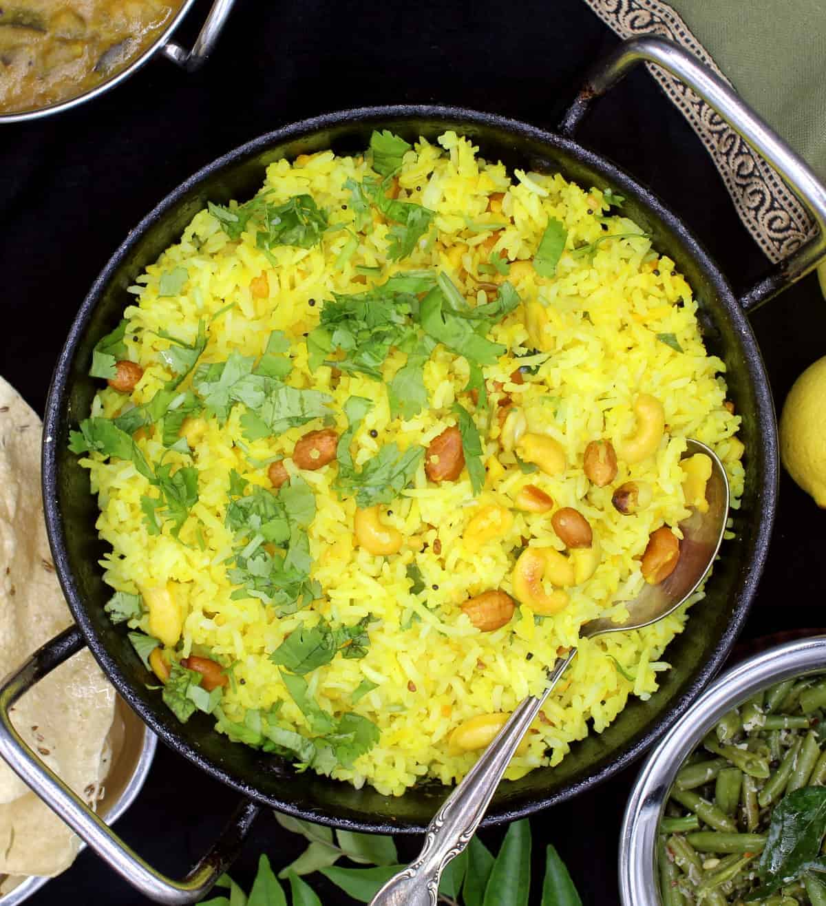 Overhead shot of a steel kadhai with bright yellow lemon rice with mustard seeds, cilantro, peanuts, cashews and red pepper, surrounded by a green bean curry, south Indian sambar and poppadum with curry leaves on the side. Also in the shot is a green and gold napkin and lemons on a black background.