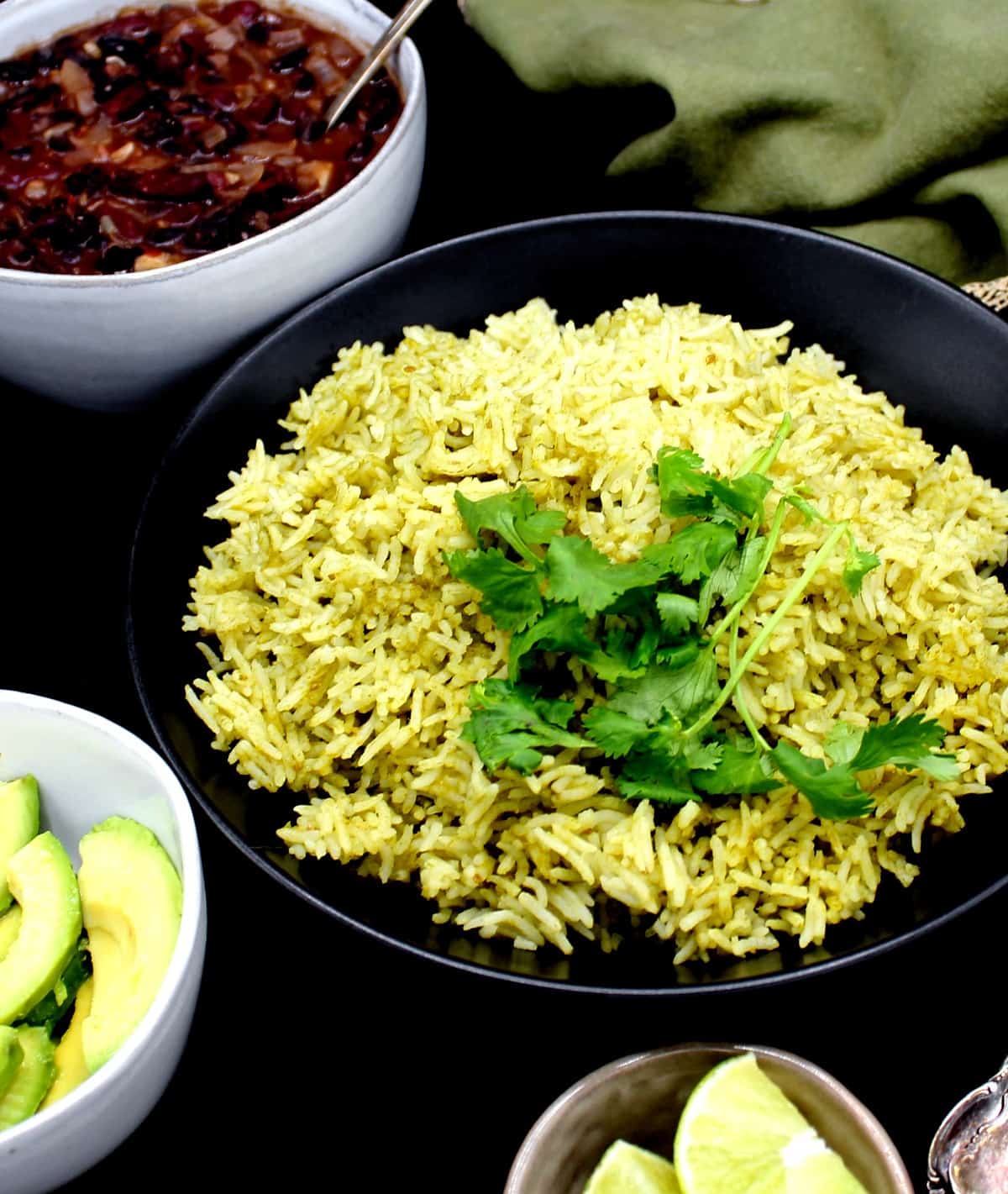 Front shot of a black bowl with Mexican green rice and cilantro, surrounded by small ceramic bowls with wedges of lime, avocado and refried beans, In the background is a green napkin with gold border, and all of it is on a black background.