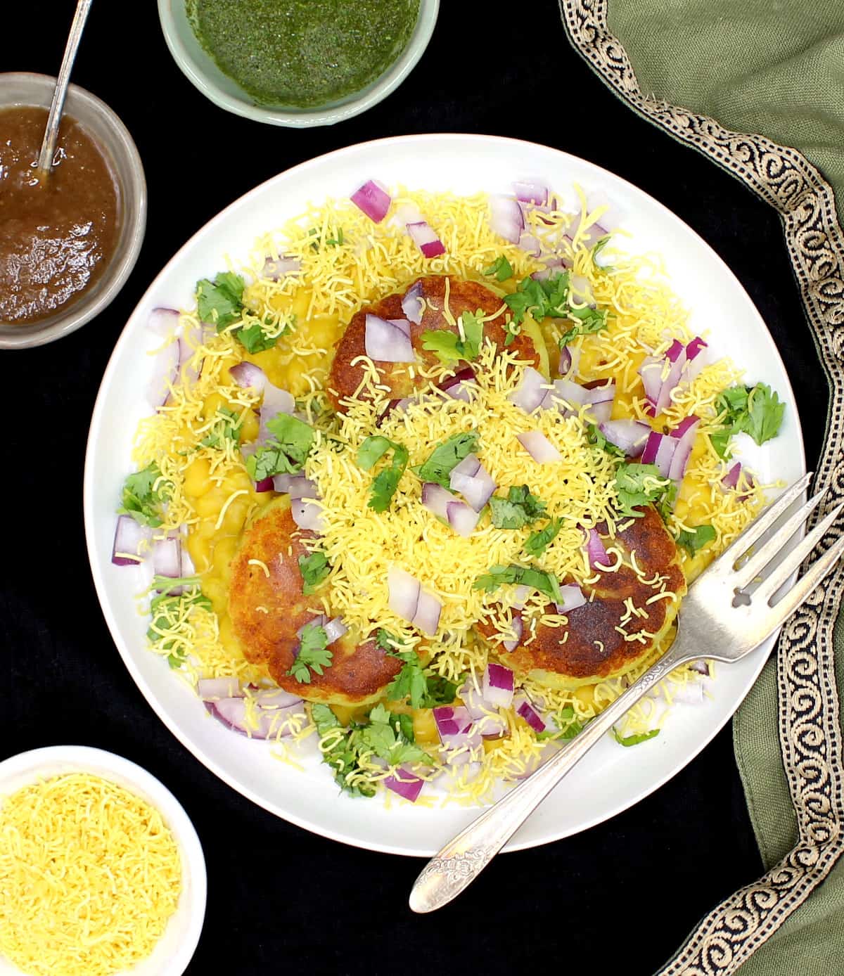 An overhead shot of a white plate of ragda pattice with potato patties, white pea sauce, tamarind-date and mint chutneys, smothered in onions, sev and cilantro.