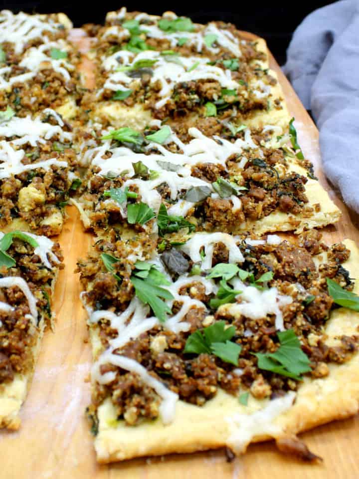 A closeup of slices of vegan breakfast sausage pizza with a biscuit crust with cheese and parsley and other herbs on a chopping board.