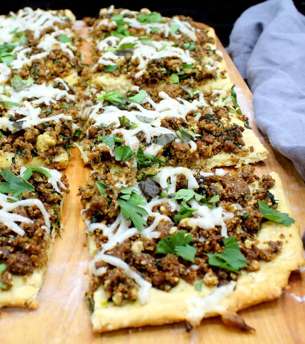 Slices of vegan breakfast sausage pizza with a biscuit crust with cheese and parsley and other herbs on a chopping board.