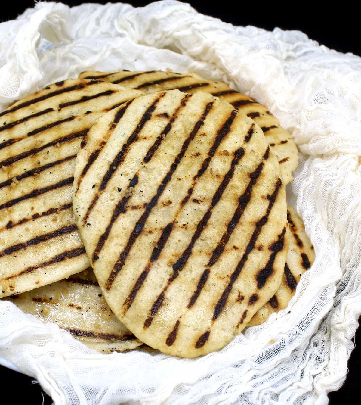 A stack of grilled vegan gluten free naans on a white plate wrapped in cheesecloth on a black background.