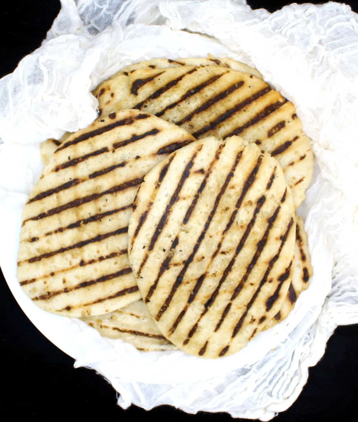 A stack of grilled vegan gluten free naans on a white plate wrapped in cheesecloth on a black background.