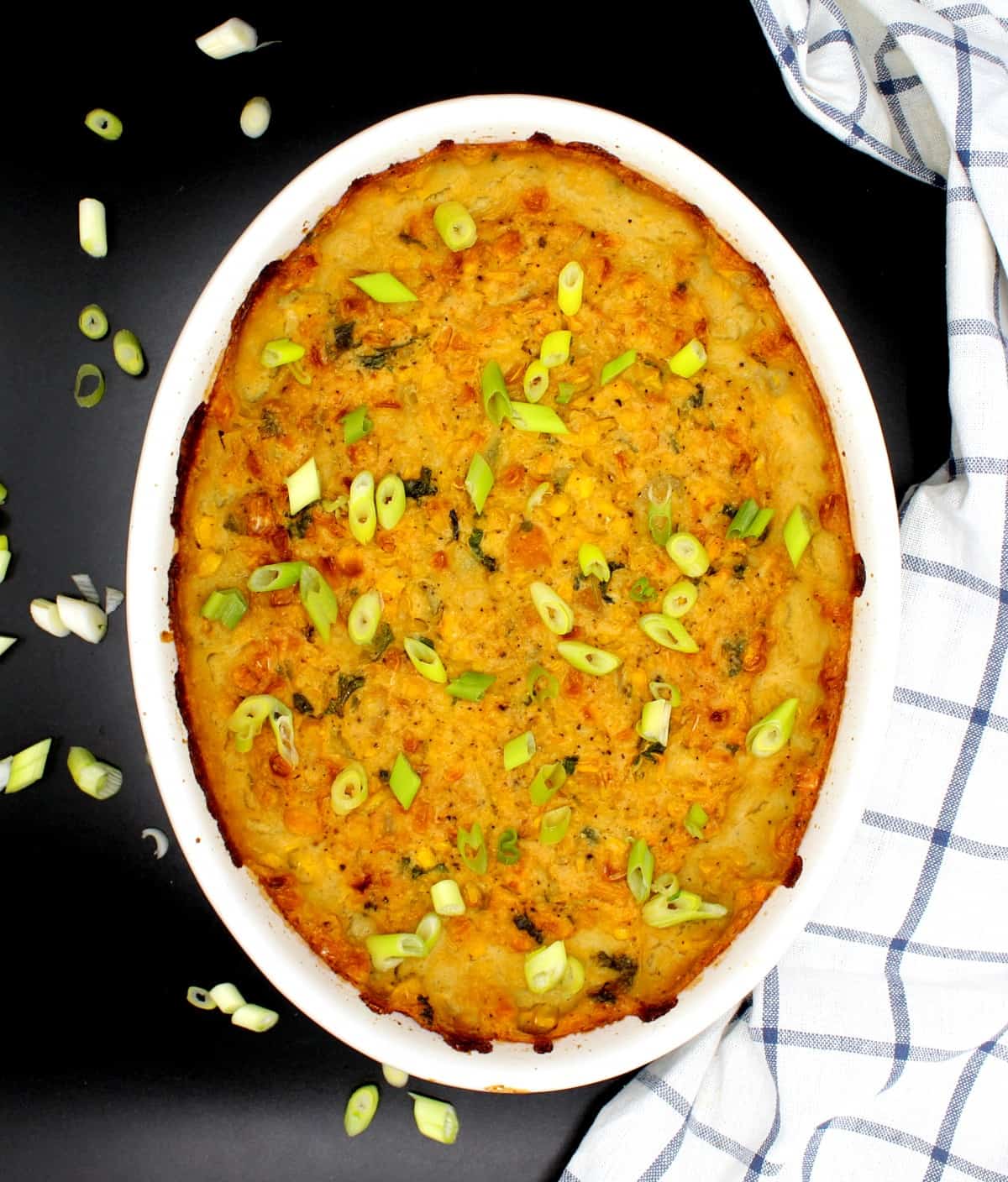 White oval casserole stoneware baking dish with savory vegan corn pudding strewn with scallions. Next to the dish is a blue and white napkin on a black background.