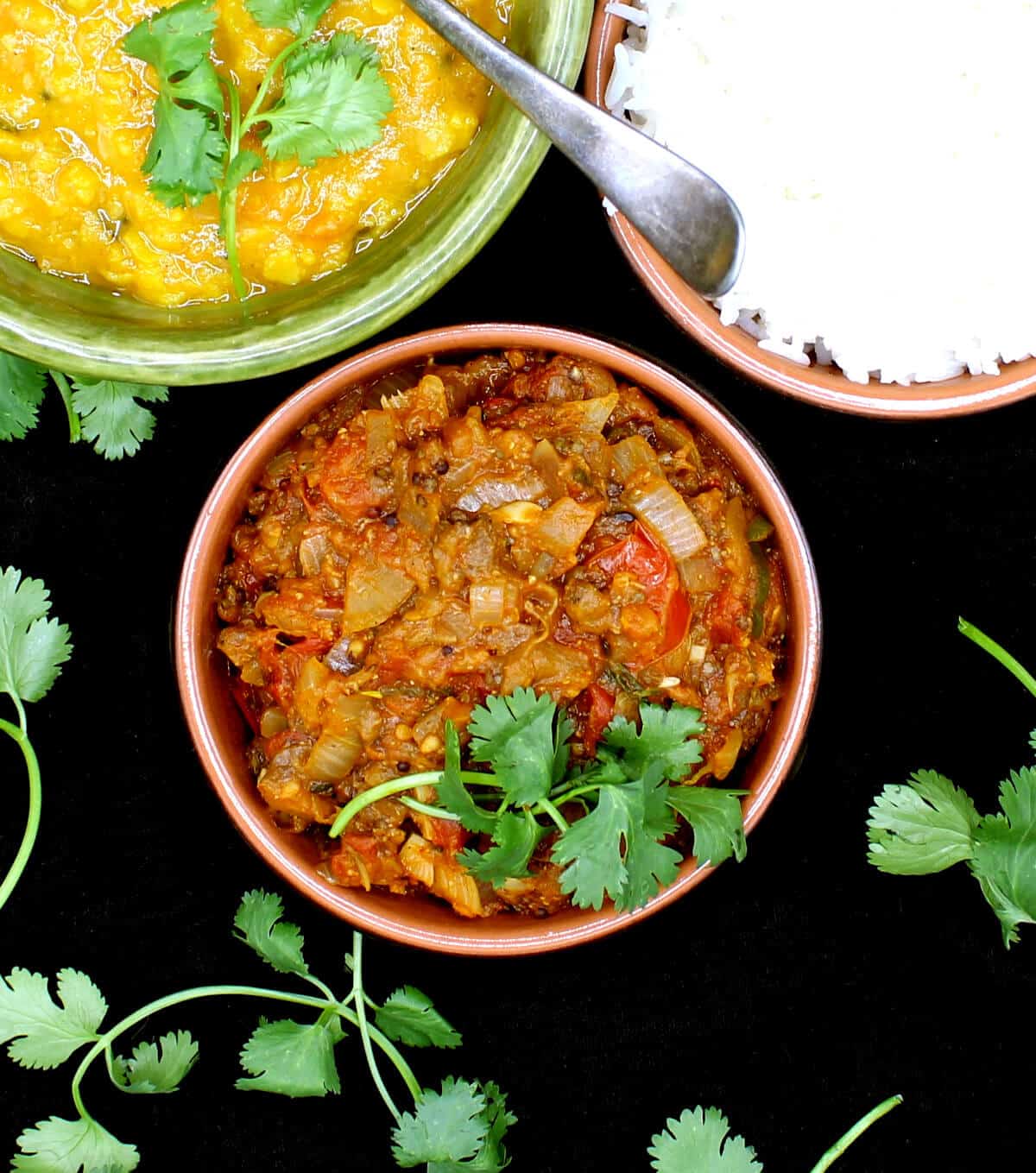 Baingan Bharta garnished with cilantro in clay bowl. Next to it are tomato dal and rice with a spoon.