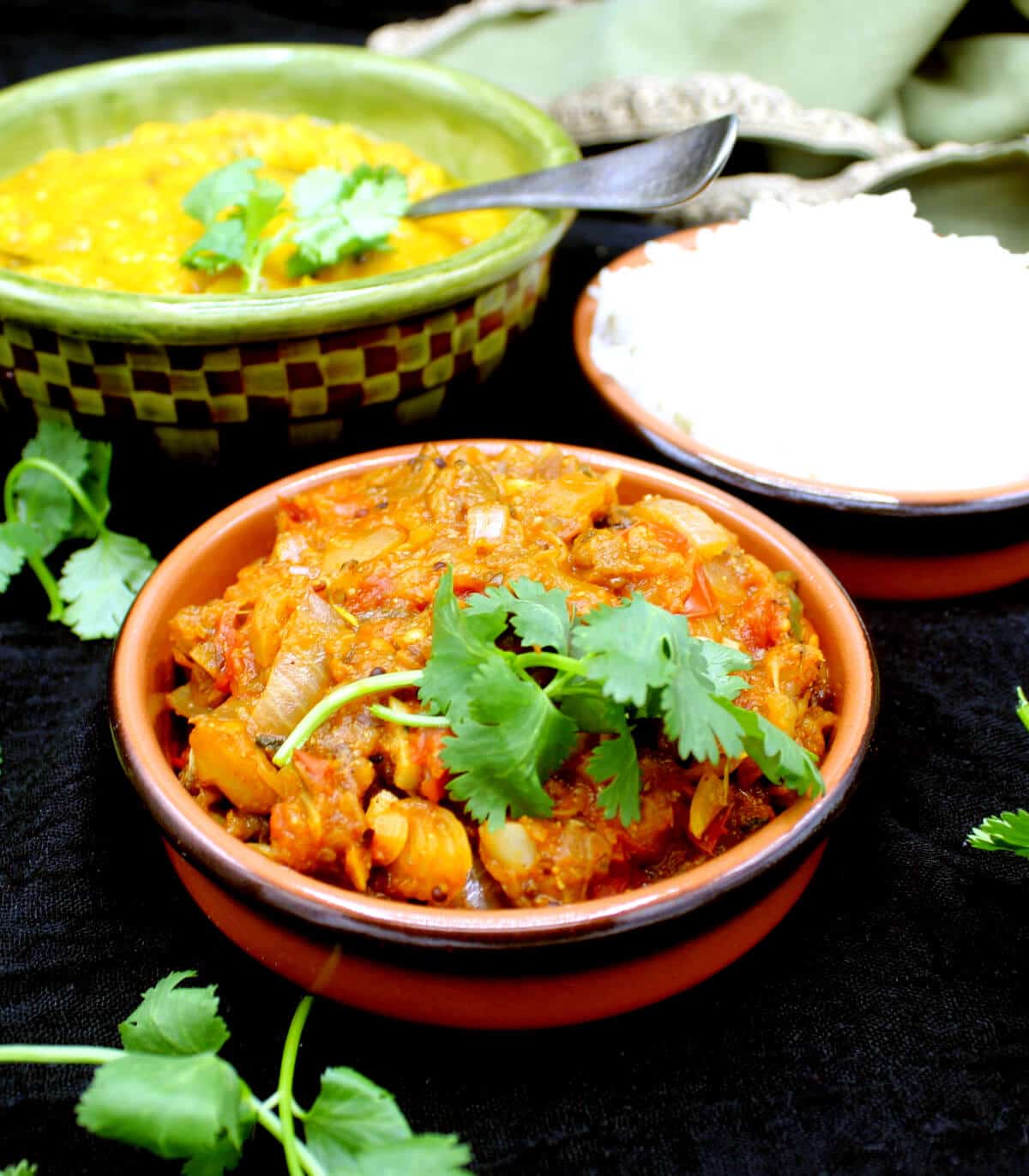 Baingan Bharta garnished with cilantro in a clay bowl. Next to it are tomato dal and rice with a spoon.