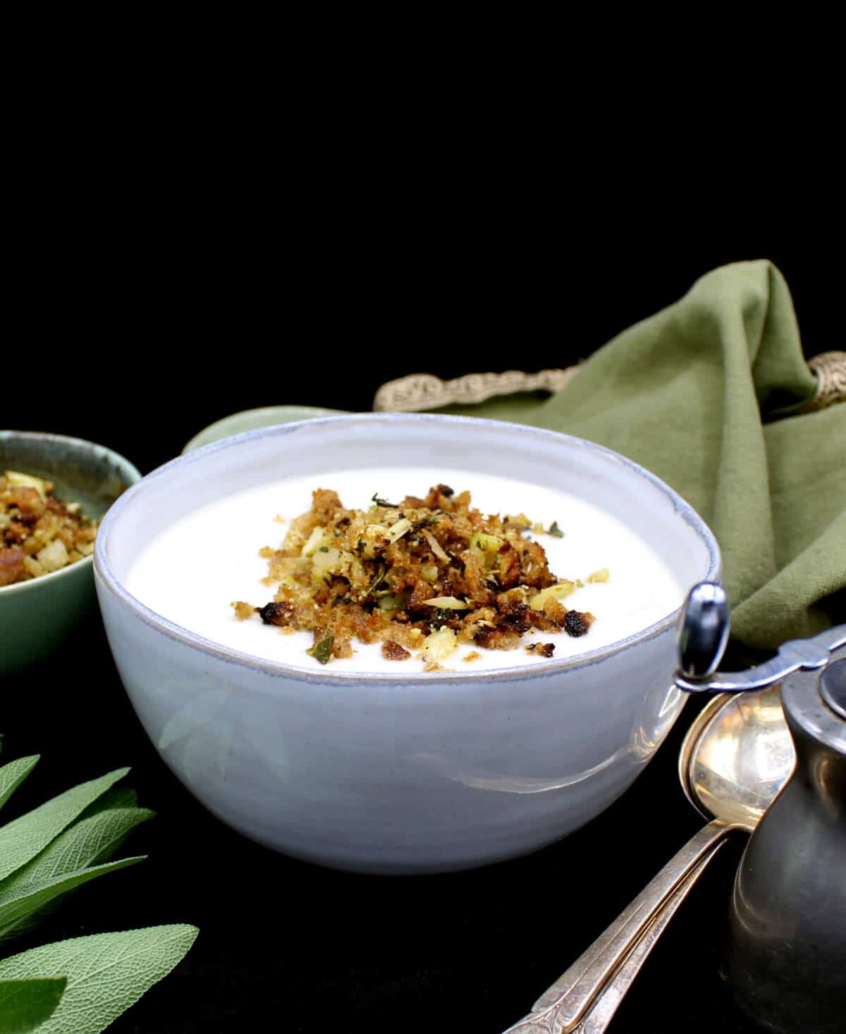 Creamy vegan cauliflower soup in a gray ceramic bowl with crunchy golden breadcrumb topping, with sage leaves, silverware, a pepper grinder and a green napkin next to it.