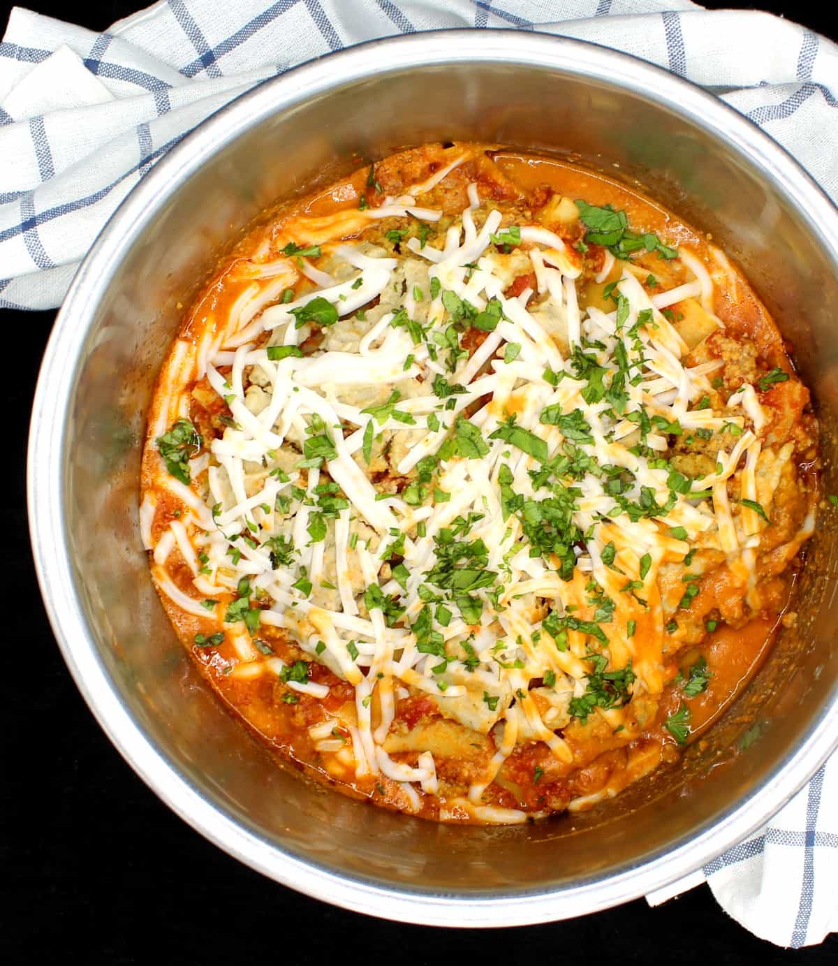 Vegan Instant Pot Lasagna with layers of creamy cashew ricotta, vegan parm and shreds of vegan mozzarella and parsley in a steel IP liner with a blue and white kitchen towel.