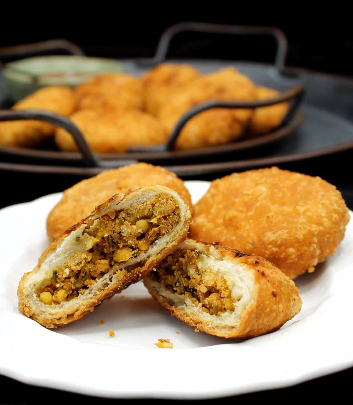 Two kachoris on a white plate with a stuffing of moong dal, besan, fennel and other spices, with a serving tray of more kachoris in the background.