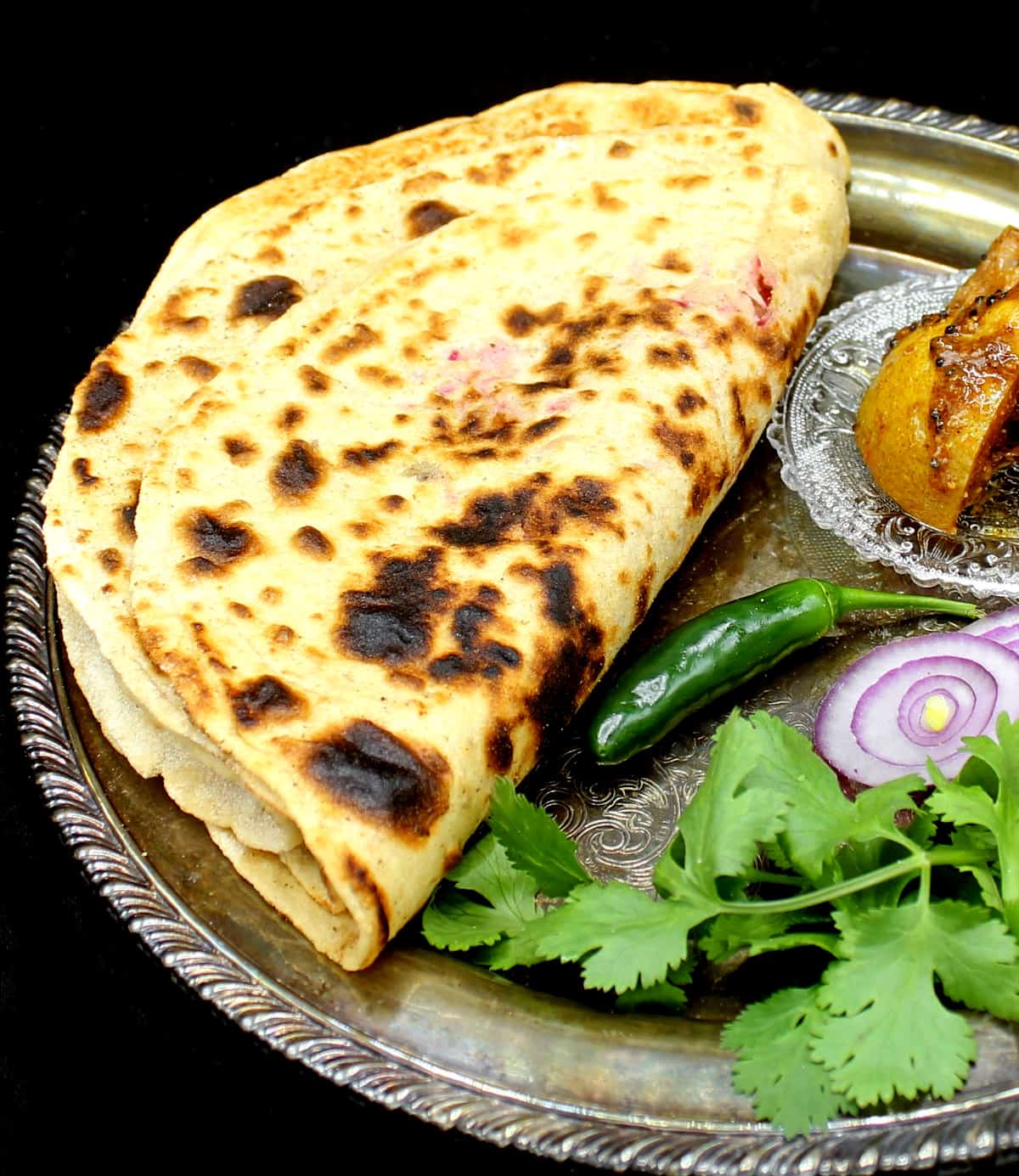 A silver decorative plate with three mooli parathas stuffed with radish and spices served alongside Indian pickles, green chili pepper, onions and cilantro