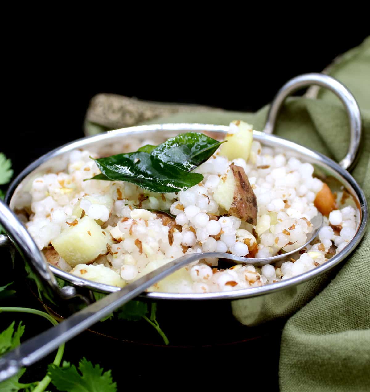 A steel wok or kadhai with sabudana khichdi, made with tapioca pearls, with a garnish of curry leaves, peanuts, potatoes and with cilantro leaves scattered around. Next to the wok is a green and gold napkin on a black background.