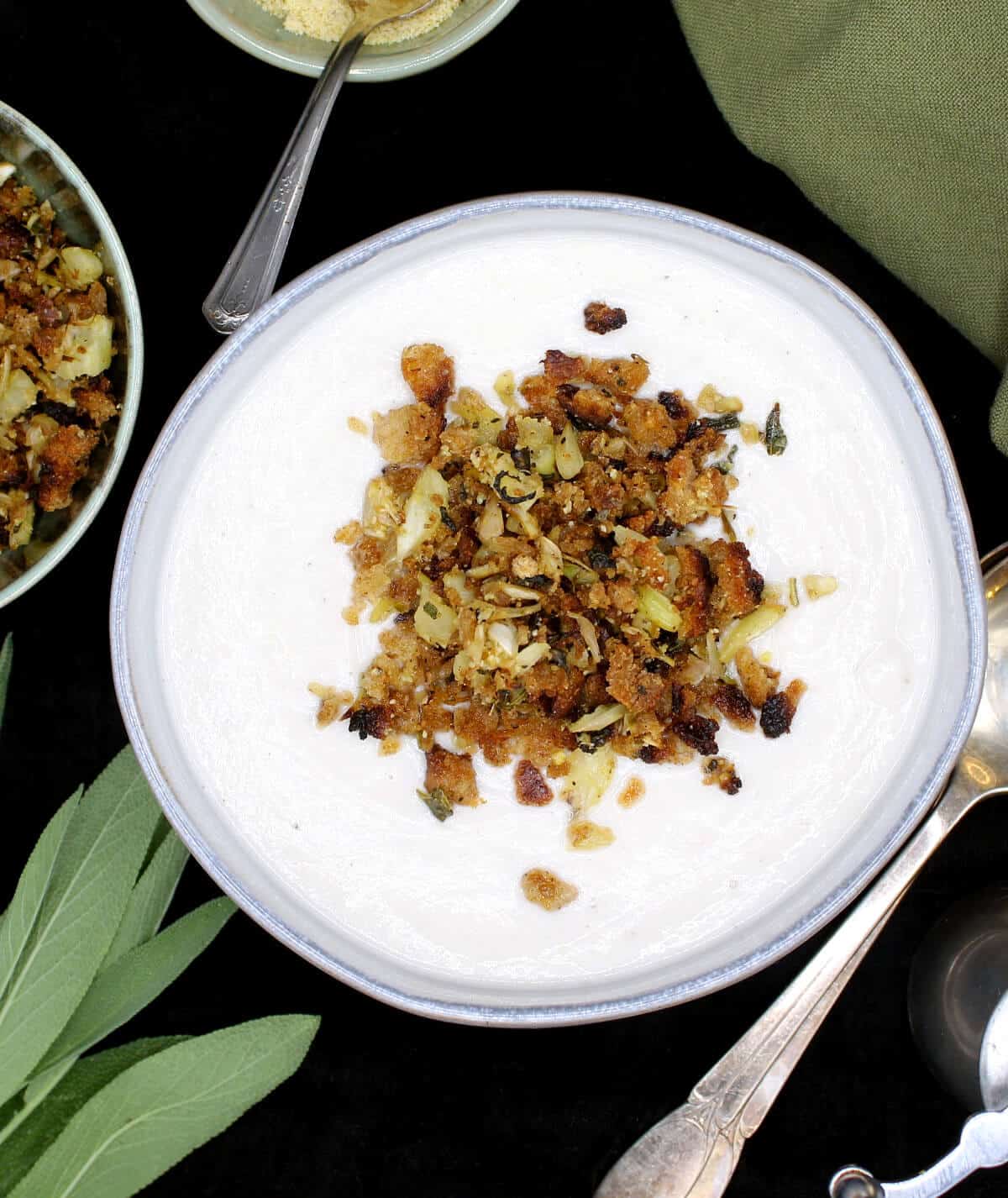 Creamy vegan cauliflower soup in a gray ceramic bowl with crunchy golden breadcrumb topping, with sage leaves, silverware, a pepper grinder, cashew parm, and a green napkin next to it.