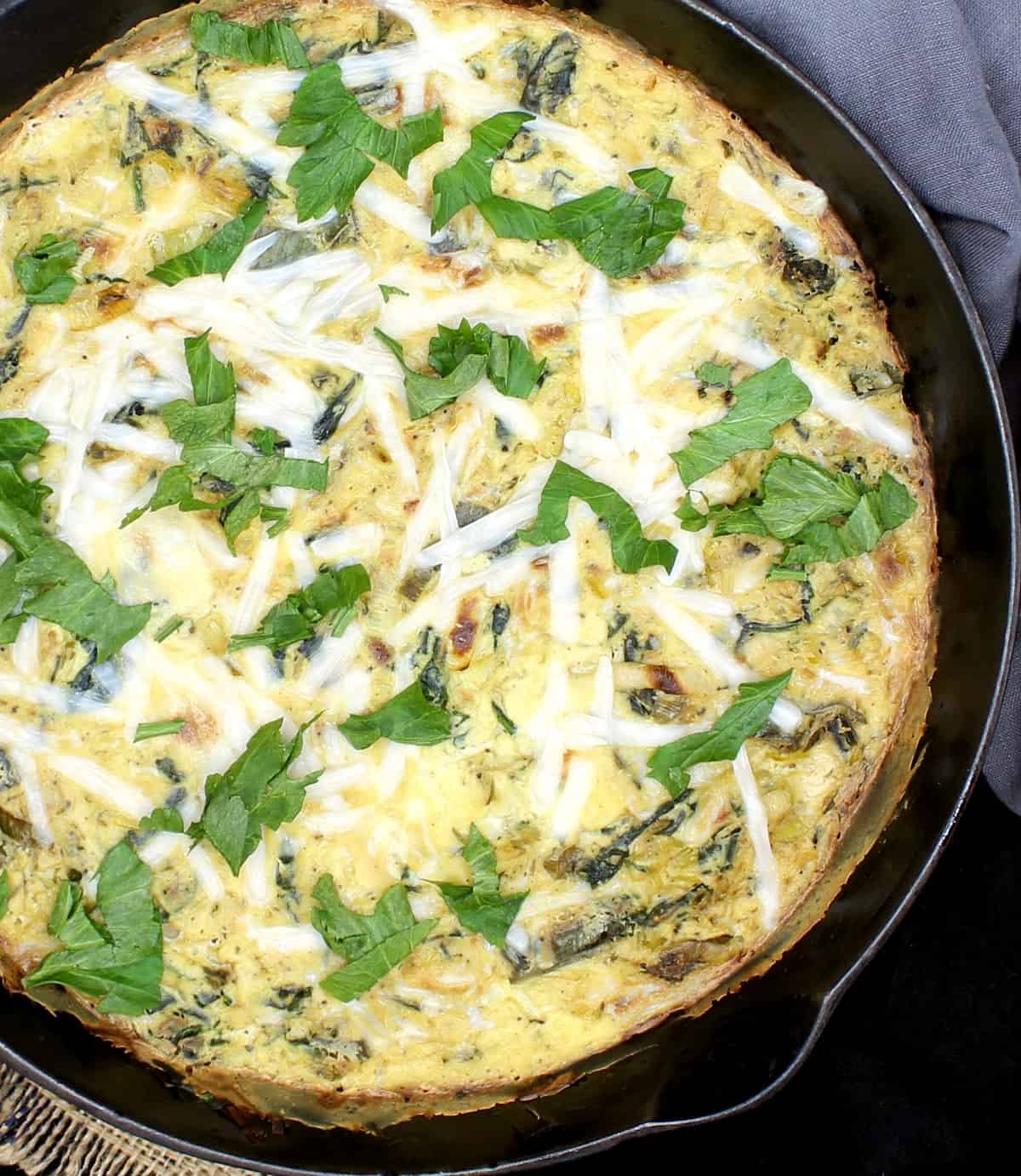 A vegan leek and spinach frittata garnished with vegan mozzarella cheese and parsley in a black cast iron skillet with a gray napkin.