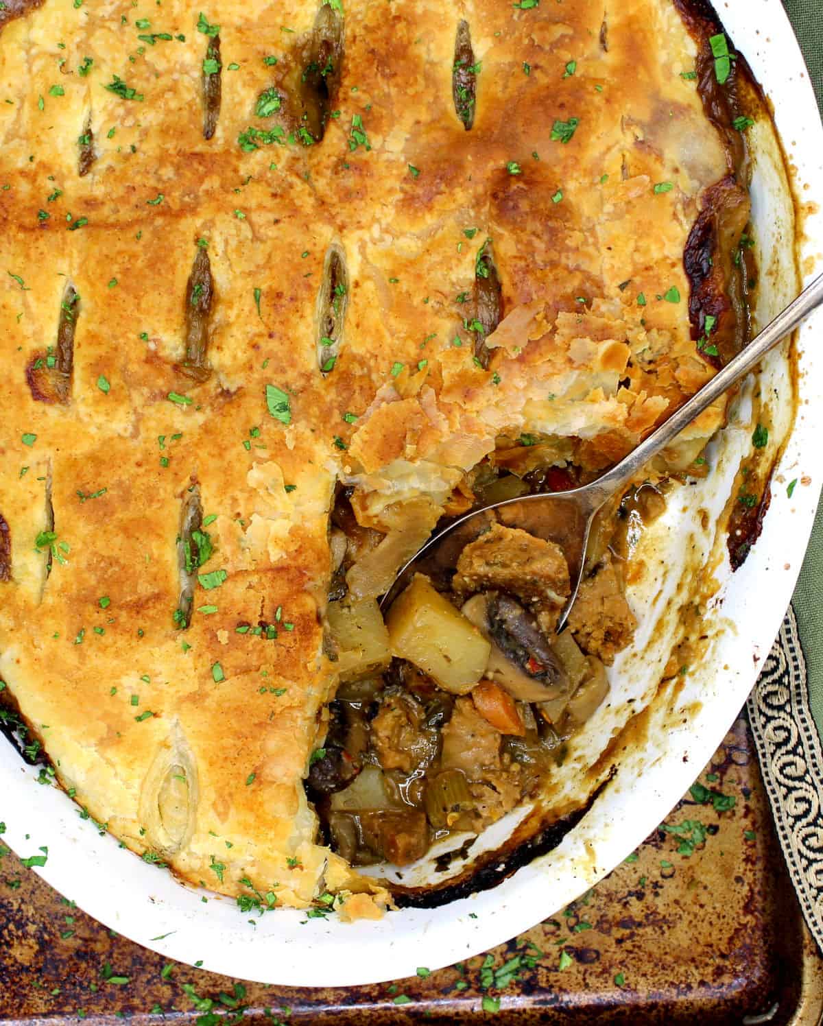An oval white baking dish with a vegan pot pie that has a flaky golden crust made of puff pastry, with a filling of potatoes, mushrooms, carrots, onions, herbs and sausage on a baking sheet with a green napkin