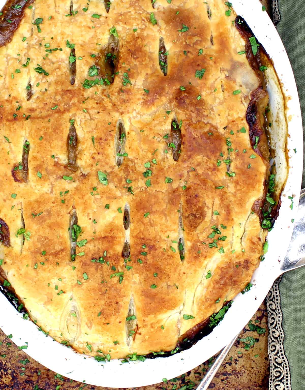 An oval white baking dish with a vegan pot pie that has a flaky golden crust made of puff pastry, with a filling of potatoes, mushrooms, carrots, onions, herbs and sausage on a baking sheet with a green napkin