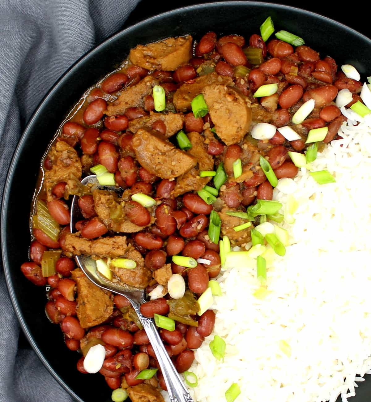 Photo of vegan red beans and rice garnished with scaliions in a black bowl with a decorative spoon against a gray napkin