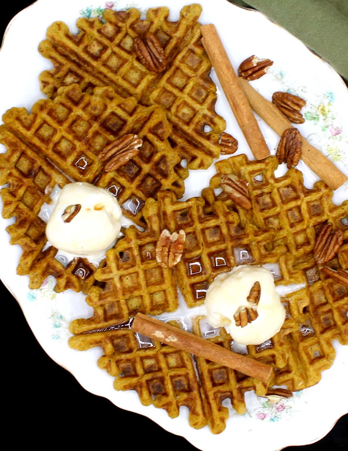 Vegan Sourdough Pumpkin Spice Waffles on a white serving dish with a flower pattern. On top of the waffles is vanilla ice cream and sticks of cinnamon and pecans are scattered around.