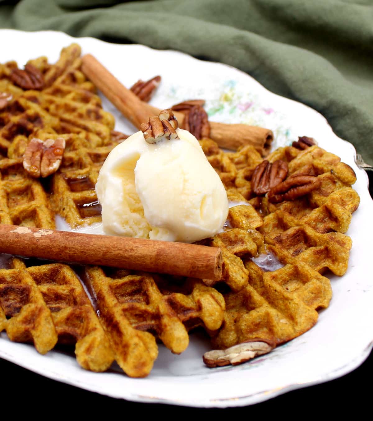 Vegan Sourdough Pumpkin Spice Waffles on a white oval decorative serving dish with a flower pattern. On top of the waffles is vanilla ice cream and sticks of cinnamon and pecans are scattered around.
