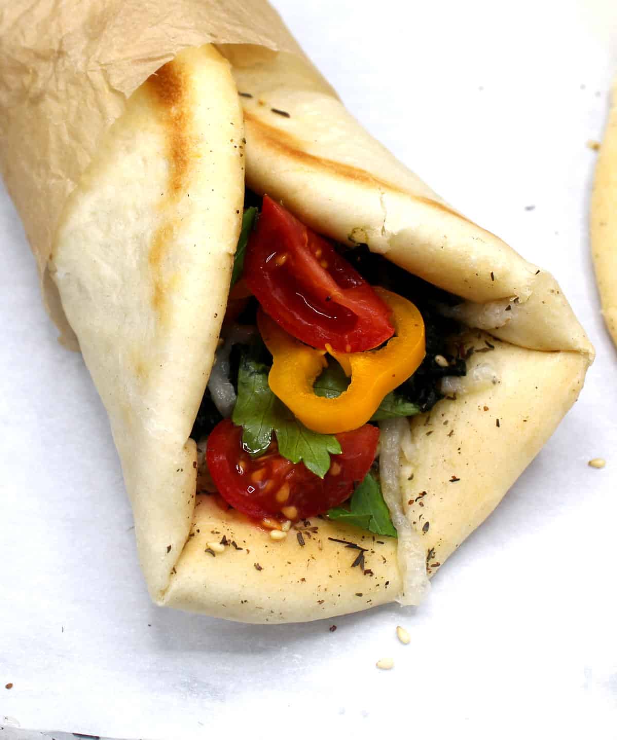 A Manakish wrap with sweet peppers, cherry tomatoes, parsley and spinach, spiced with cumin, coriander and za'atar, rolled up into a wrap and secured with brown parchment paper.
