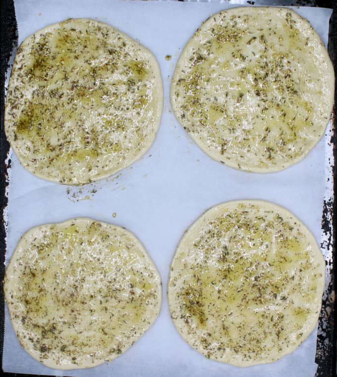 Four unbaked manakish flatbreads on a parchment paper on a baking sheet. Flatbreads are smeared with olive oil and za'atar mixture