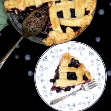 A golden, delicious slice of blueberry pie on a blue and white plate with an intricate design and a fork with the full pie next to it