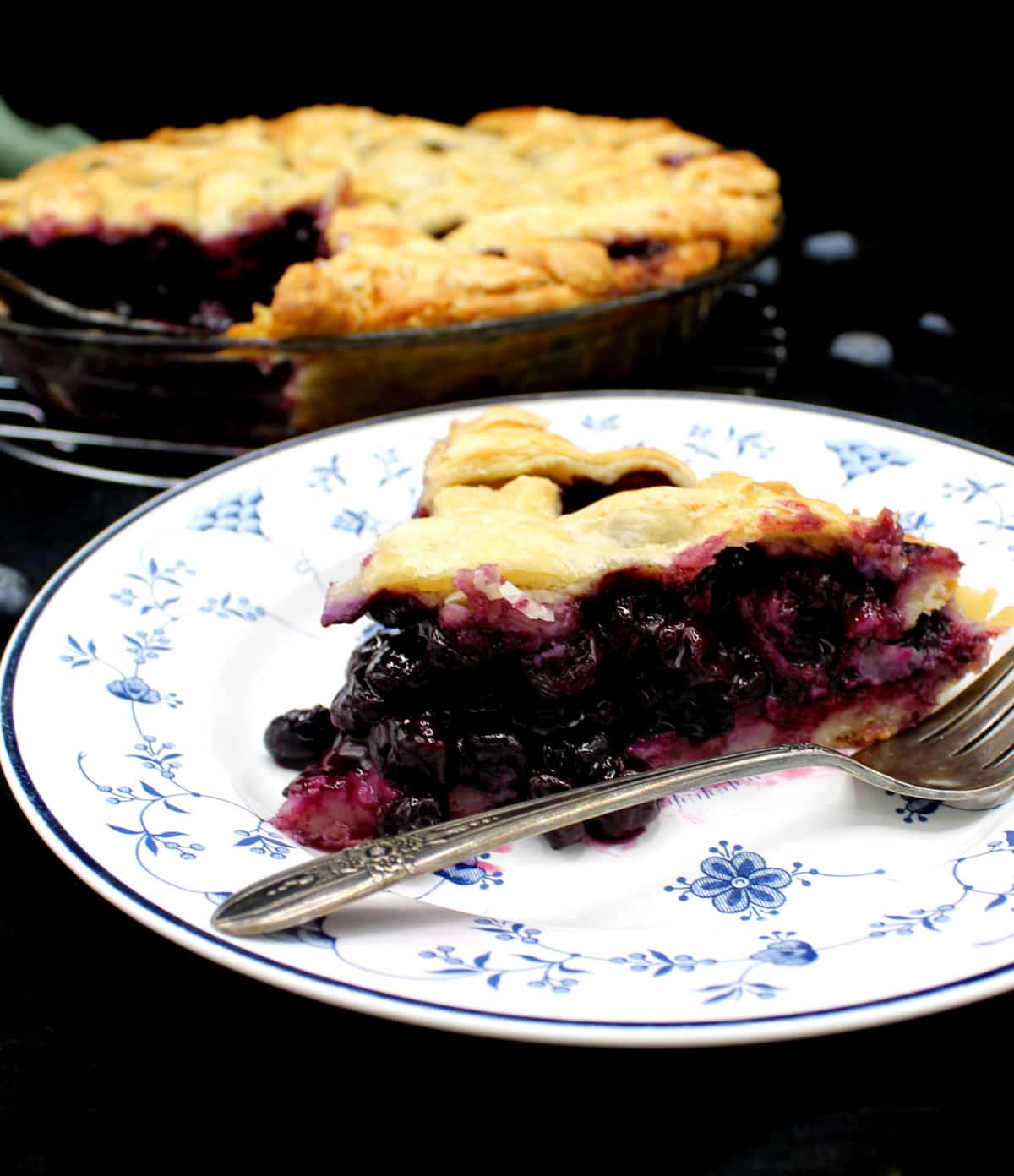 A slice of vegan blueberry pie on a blue and white china plate with a delicate floral pattern and a silver fork with the whole pie next to it and blueberries scattered around.