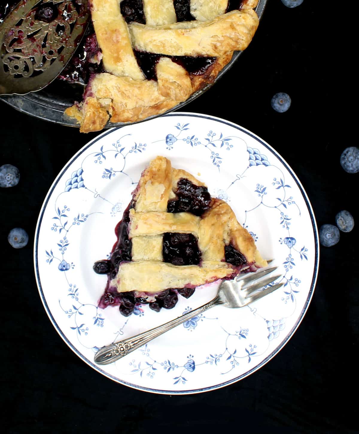 A slice of vegan blueberry pie on a blue and white china plate with a delicate floral pattern and a silver fork with the whole pie next to it and blueberries scattered around.