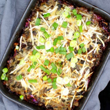 A stoneware gray baking dish with a vegan cabbage casserole with meaty vegan beef crumbles, cabbage, potatoes, herbs and vegan mozzarella shreds and scallions scattered on top, on a gray napkin