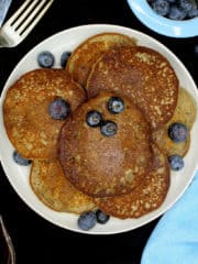 An overhead shot of a plate filled with vegan glutenfree coconut flour pancakes with blueberries and next to it are a bowl of blueberries, a blue napkin, a kinfe and fork.