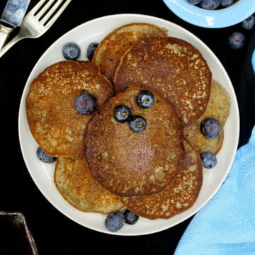 An overhead shot of a plate filled with vegan glutenfree coconut flour pancakes with blueberries and next to it are a bowl of blueberries, a blue napkin, a kinfe and fork.