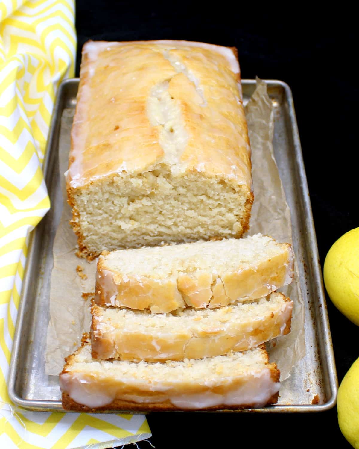A loaf of buttery vegan lemon pound cake on a metal baking tray with slices cut in front of it and showing the dense, soft crumb. Next to it are lemons and a yellow and white napkin.