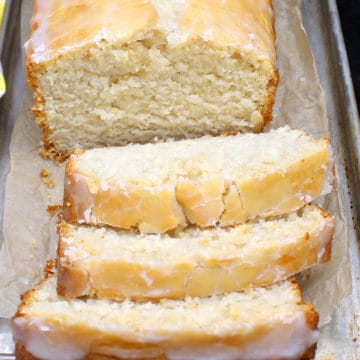 A loaf of vegan lemon pound cake with three slices cut and showing the dense, soft crumb on a metal pan with lemons and a yellow and white napkin
