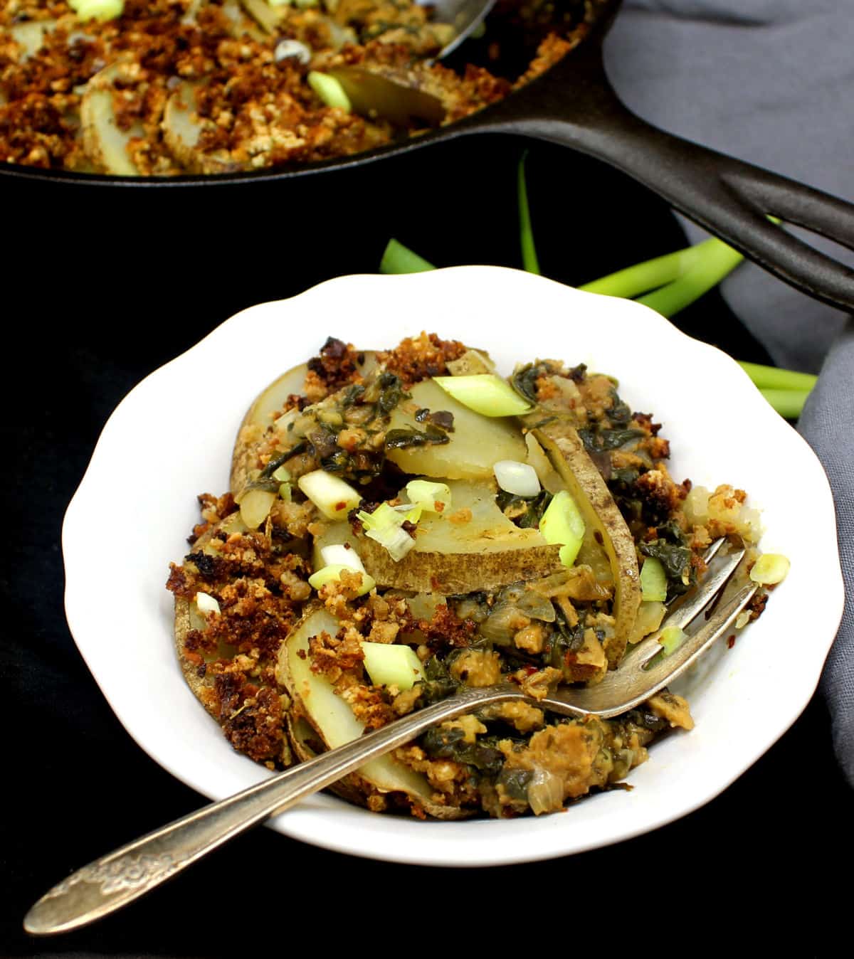 A white ceramic bowl with a serving of vegan spinach potato casserole with sausage and a fork. Scallions are scattered on top and on the side.