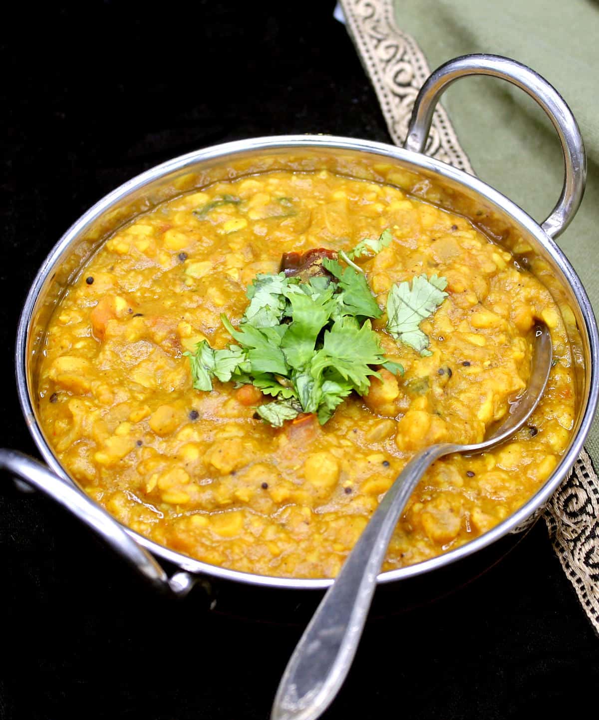 A closeup of a steel and copper kadhai with panchmel dal and cilantro with a green and gold napkin.