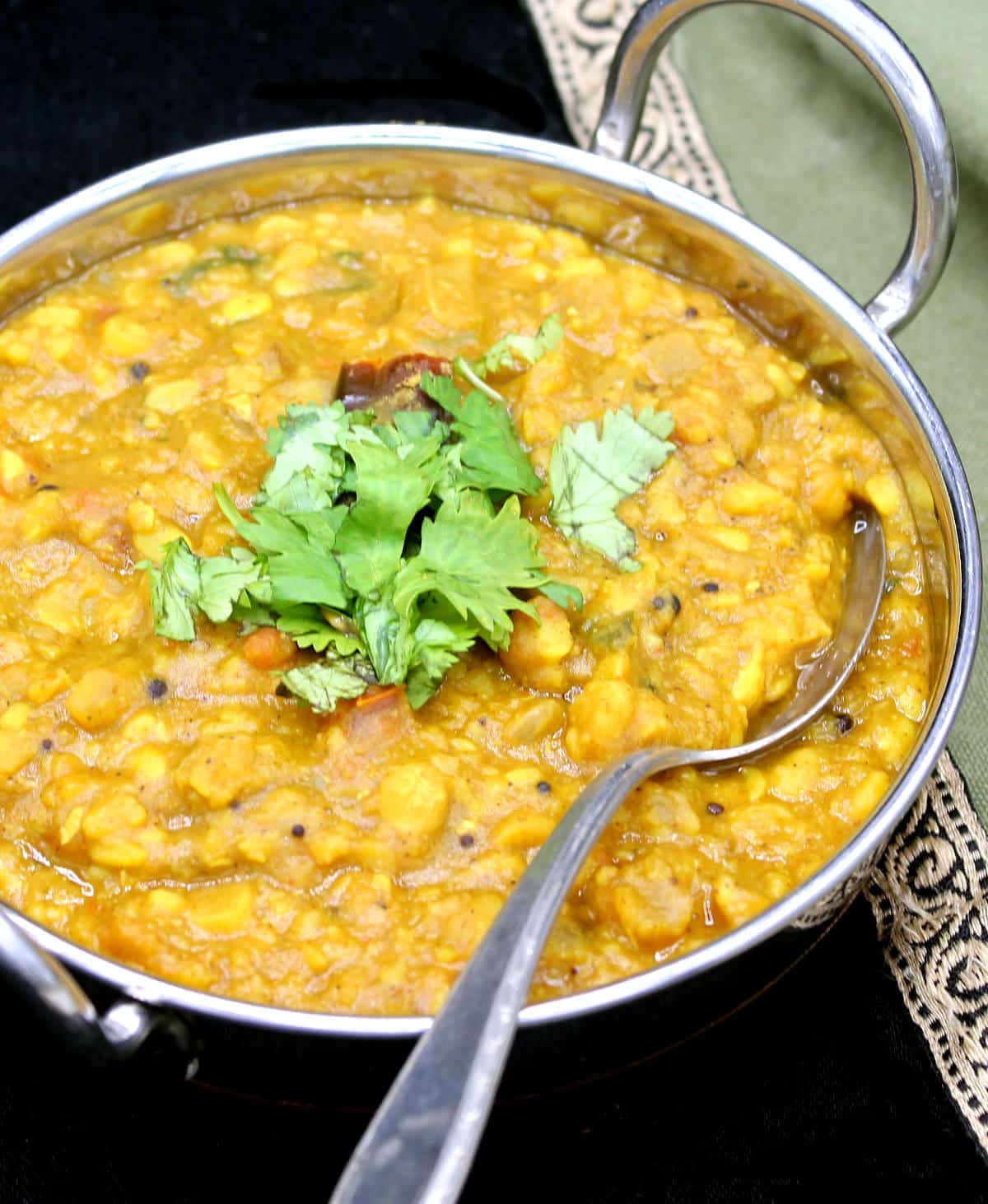 Closeup of dal with coriander leaves in a steel kadhai with a silver spoon and a napkin in green with gold border.