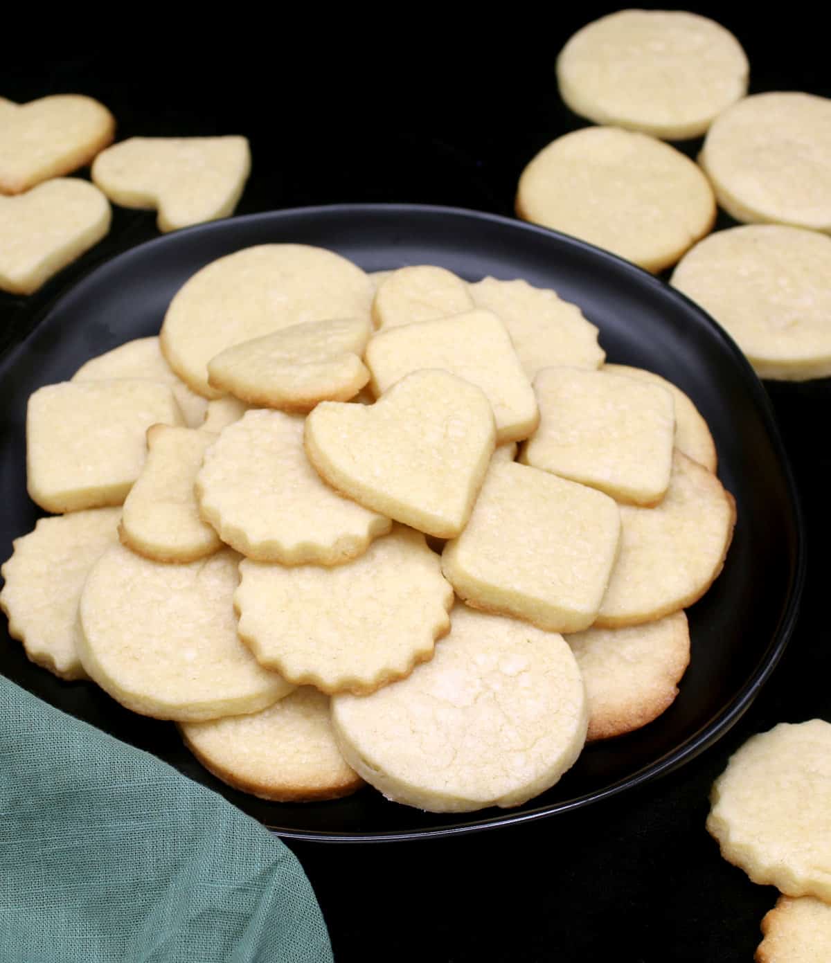 A closeup of vegan sugar cookies inside and around a black plate, cut out in discs, hearts and squares.