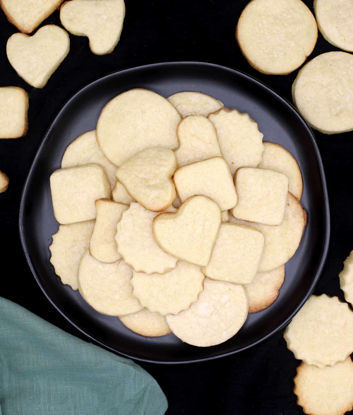 Vegan sugar cookies inside and around a black plate, cut out in discs, hearts and squares.