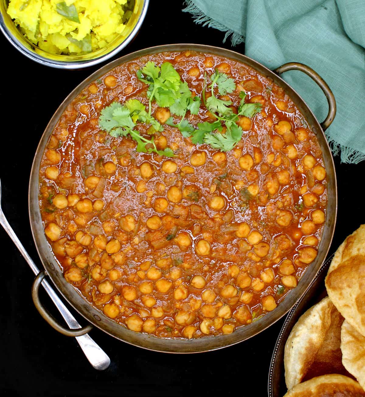 Overhead shot of chana masala in copper server with pooris and potato sabzi on the side and a serving spoon.