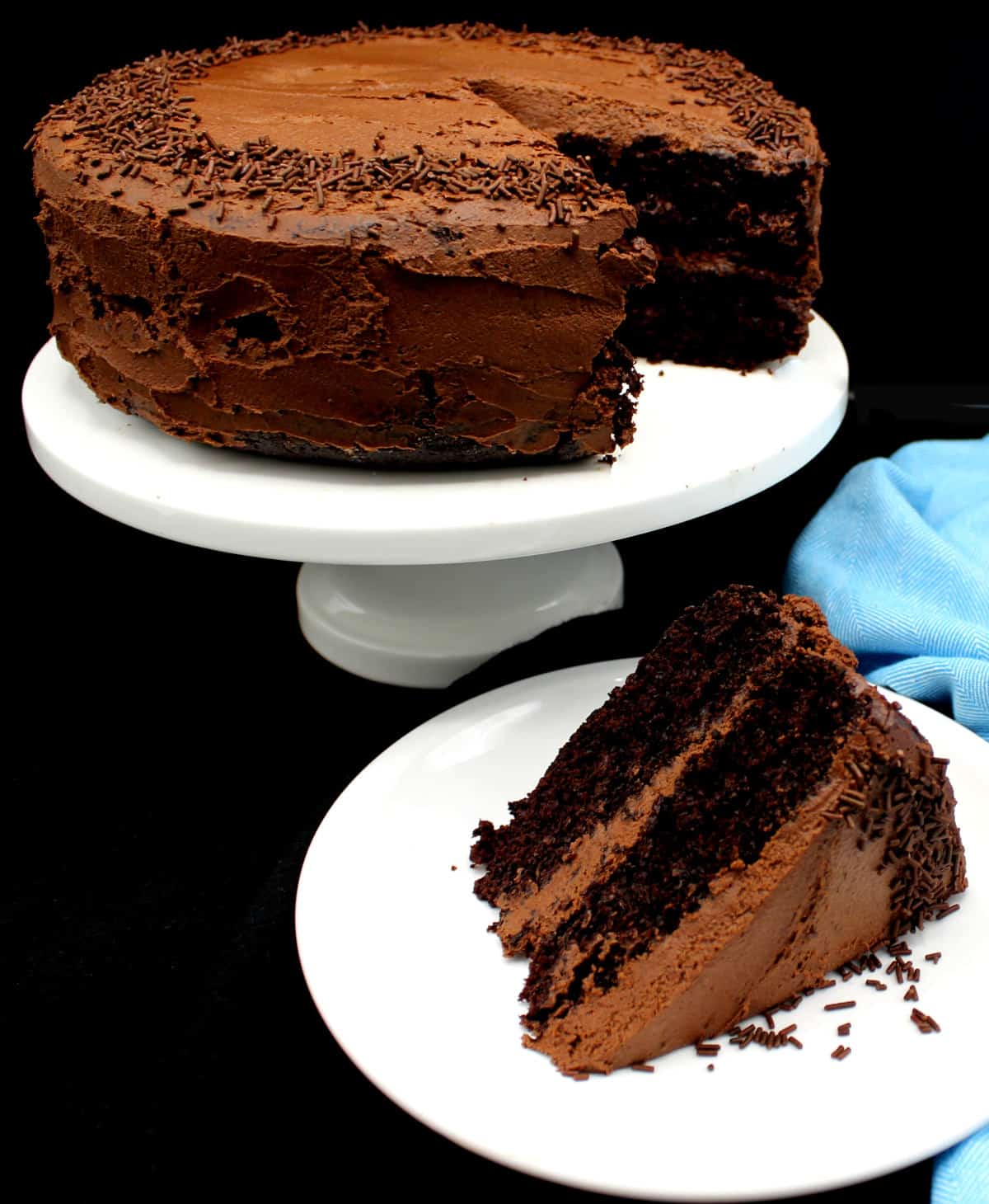 A slice of a two-layer vegan chocolate cake on white plate with full cake on cake stand in background.