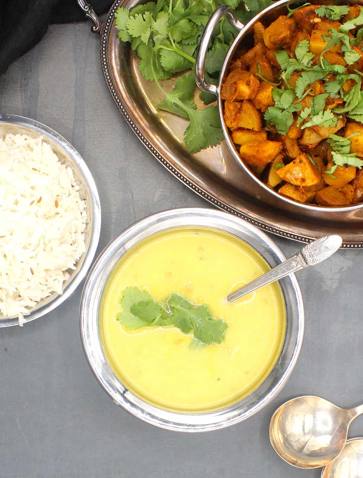 Creamy moong dal in a copper and steel serving bowl with a silver spoon and rice and Bombay potato sabzi on the side.