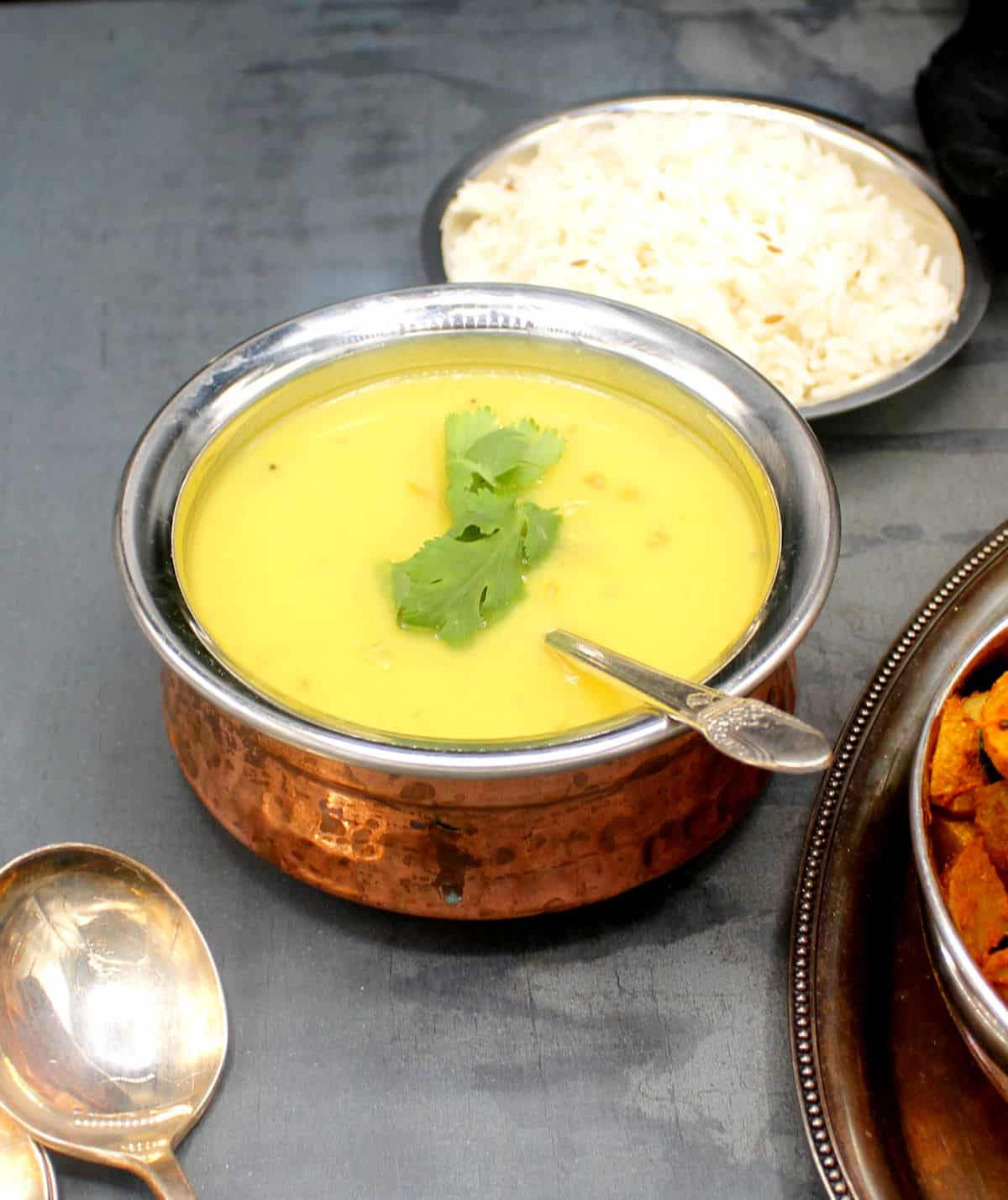 Creamy moong dal in a copper and steel serving bowl with a silver spoon and rice and Bombay potato sabzi on the side.