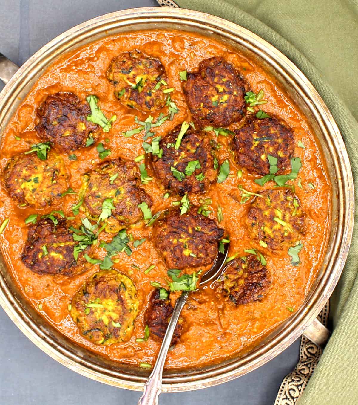 Overhead closeup of zucchini kofta curry--kofta balls in a spicy orange sauce in a silver serving dish with a spoon and green napkin.