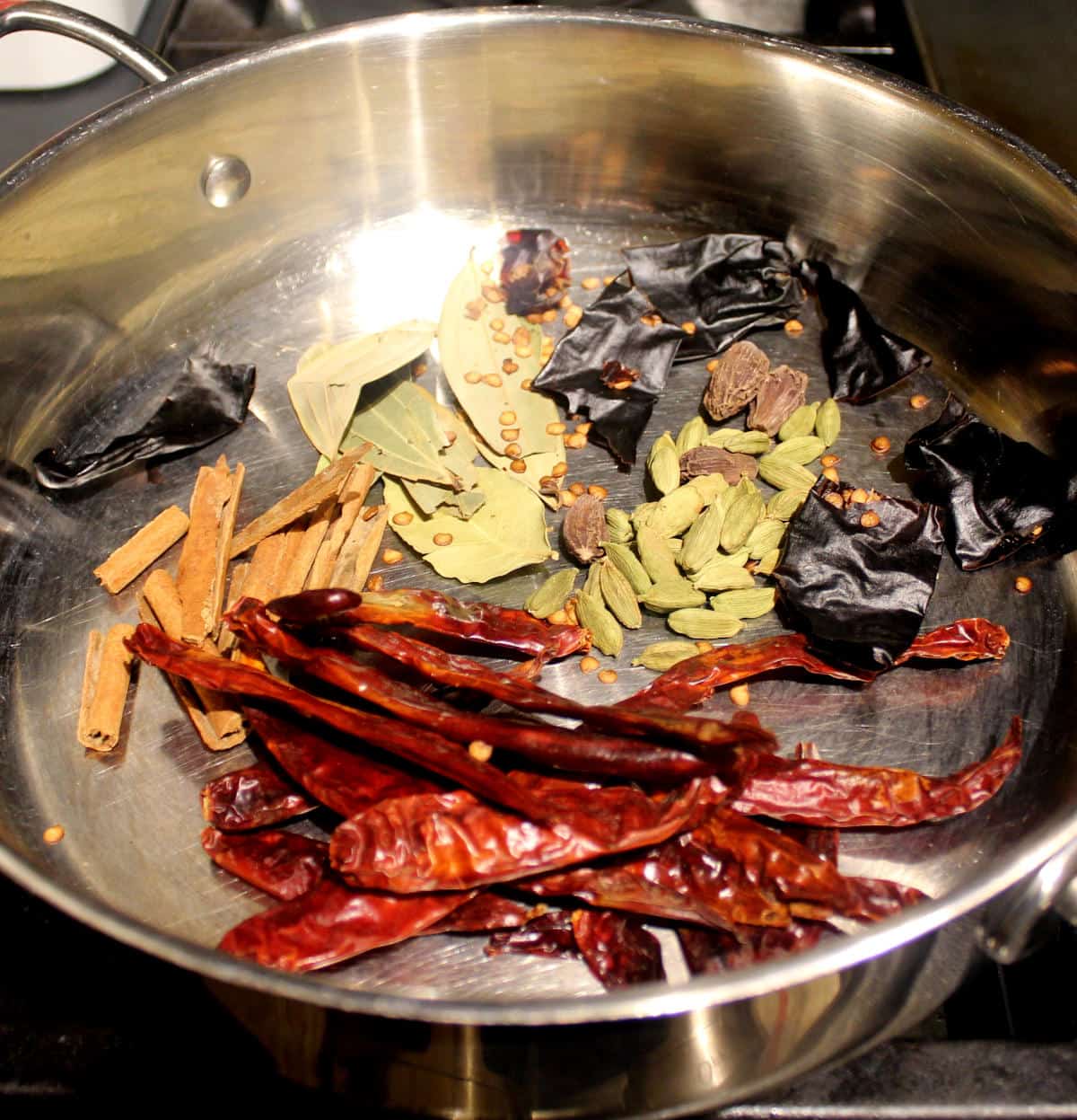 Large spices for garam masala, including bay leaves, chili peppers, cinnamon, cardamom, added to a wide steel skillet.
