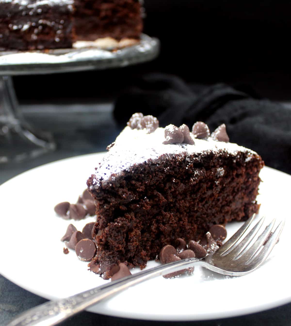 A slice of Irish chocolate whiskey cake on a plate with chocolate chips and a fork with the full cake in background on a glass cake stand.