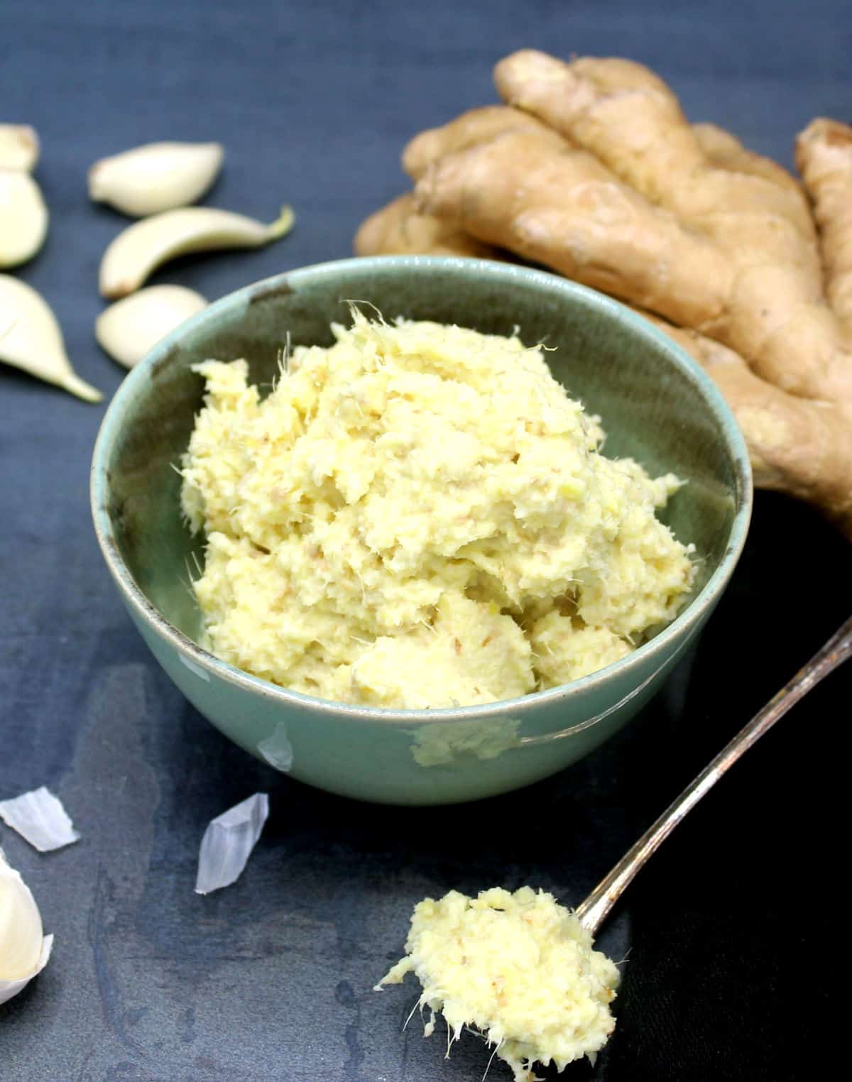 Photo of a green bowl with ginger garlic paste with gingerroot and peeled and unpeeled garlic around it.
