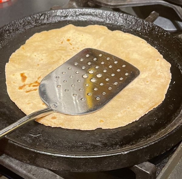 Sourdough roti browning on cast iron griddle with spatula.
