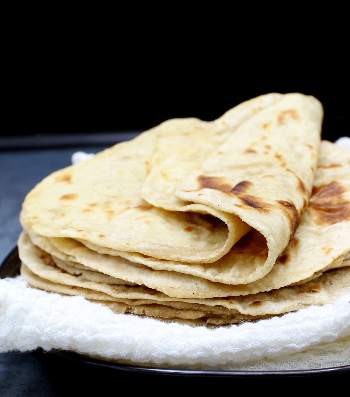 Front shot of soft sourdough rotis on a black plate wrapped in white cheesecloth.