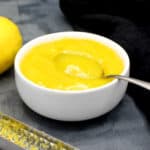Photo of lemon curd in a white bowl with a spoon and a lemon in the background as well as a zester.