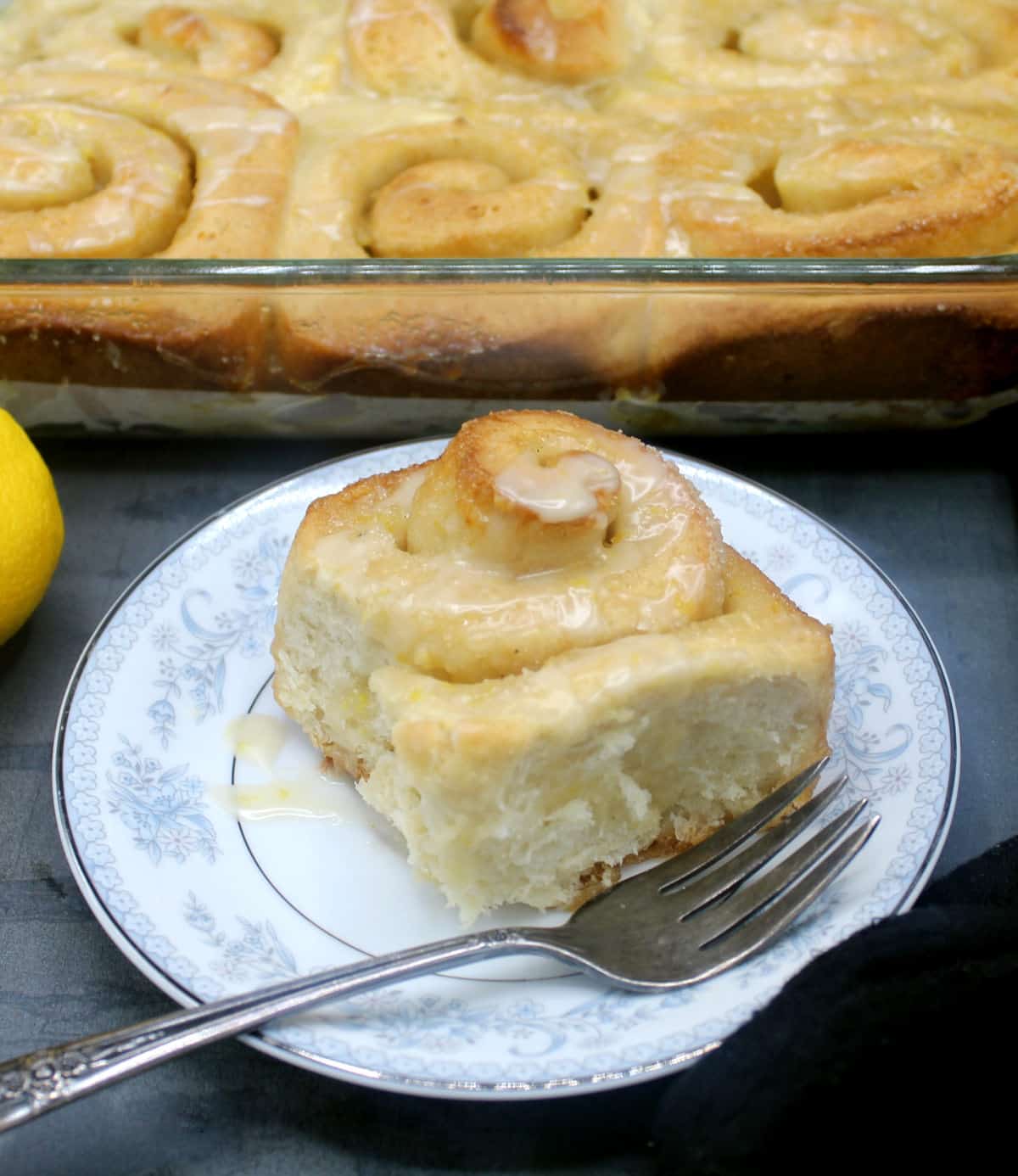 A vegan lemon sweet roll with sticky lemon glaze on a blue and white china plate with rolls in the baking dish and a lemon in the background.
