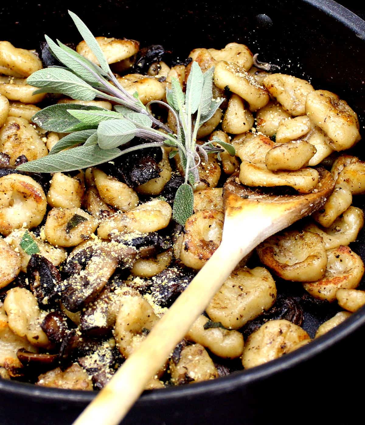 A closeup of vegan sage butter gnocchi in a skillet with sage, mushrooms and a ladle.